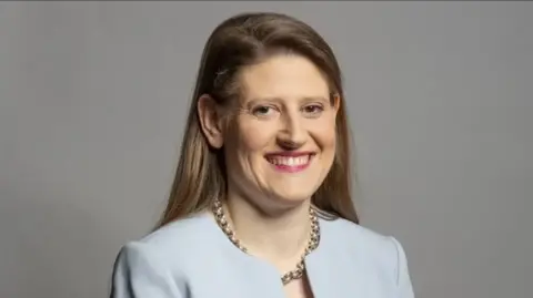 Head-and-shoulders studio portrait of a woman against a plain light grey background. The person has long brown hair parted to one side, visible teeth in a broad smile, and is looking toward the camera. The person is wearing a light blue jacket or blazer-style top and a chunky silver chain necklace. Soft, even lighting creates a professional corporate headshot appearance.
