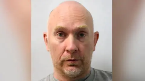 Met Police Close-up head-and-shoulders portrait of a bald Wayne Couzens with light facial stubble wearing a grey shirt, facing the camera against a plain light-coloured background. The image is tightly cropped and resembles an identification or mugshot-style photograph.