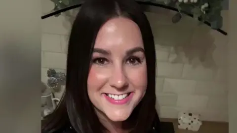 A smiling Lucy Connolly looking directly down the camera. She has shoulder length, dark brown hair and brown eyes. She is seen indoors, in what appears to be a kitchen, with a white tiled splashback and work surface behind her. The edge of a hob extractor fan can also be seen behind her.