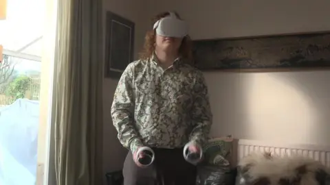 A 16 year old boy wearing a green vintage shirt with long curly hair has a virtual reality headset on and is holding two VR controls. He is standing in the living room of his family home. 
