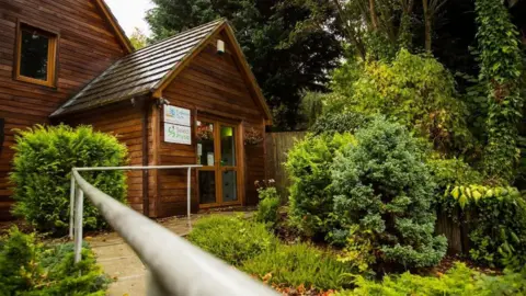 Phil Newbury A wooden building, surrounded by trees and bushes. Next to the door is a sign reading "Exclusive Gym" and another reading "Select Physio".