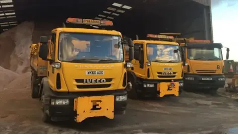 Three yellow gritting lorries wait at a depot as they are loaded up with gritting salt before heading out on the roads.