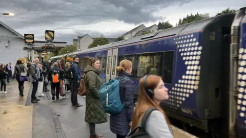 PA Media Crowd of train passengers stand on platform with blue scotrail train at station under a grey cloudy sky.