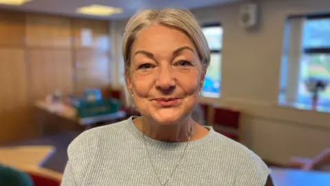 BBC A women with blonde hair tied up, she is wearing a grey ribbed jumper whilst looking at the camera smiling.