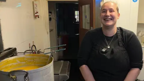 On the left of the shop is an industrial pan full of soup, with a ladle hanging in there, and on the right is Sam, wearing black clothes and a black apron. She is stood in the takeaway's kitchen.