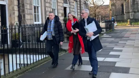 David Toshack on the right of a group of three people wearing a dark suit with a dark jacket.
