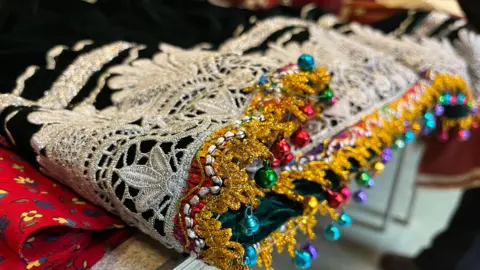 The edge of a green velvet dress, which has silver and gold lacing with bells on the bottom