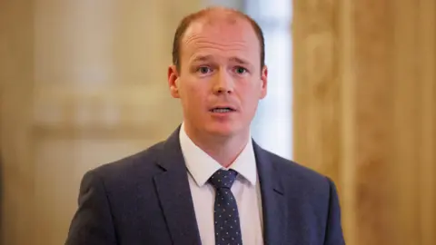 PA Media Gordon Lyons, with short brown hair, wearing a white shirt, navy tie and navy suit. 