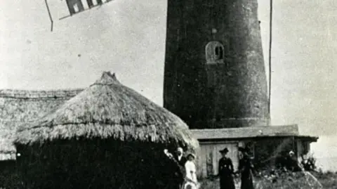 Wilton Windmill Society A black and white saturated image of the Wilton Windmill in the early 1900s with three people standing in front of it, one wearing white and the other two in long black dresses and bonnet hats. There is a small thatched building in the foreground. 