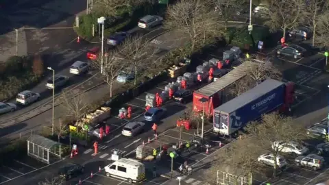 Aerial look at car park with queue of cars and people in red outfites lined up by pallets of water.