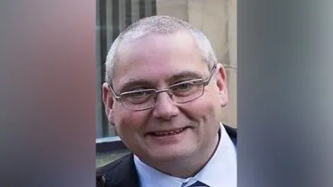 Lancashire Police David Daintree smiles into the camera. He has short grey hair and glasses and is wearing a shirt and tie and suit. 