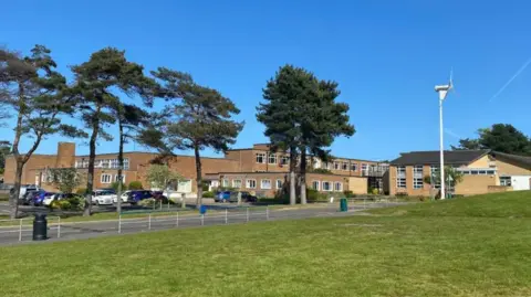 View of the Sheringham High School Building which has six tall trees in front of it. The building is sand-coloured with flat roofs. 