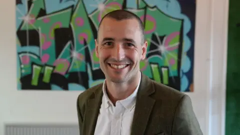 Ben Drabble has short hair and is wearing a green blazer over a white shirt. Behind him is a graffiti-style mural.