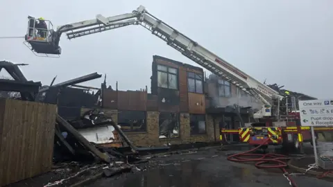 Firefighters on an aerial ladder platform douse water on a school which has burnt down. The damage to the building is clear to see which large sections of it burnt away and charred marks on the building.