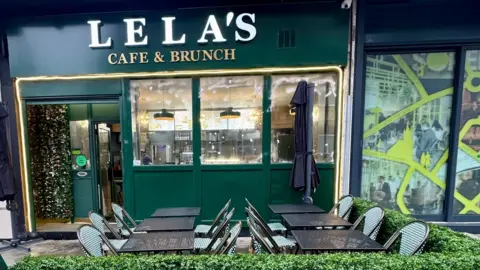 Bradford Council Cafe tables and chairs outside a green, gold and white cafe front. They are surrounded by a green hedge.