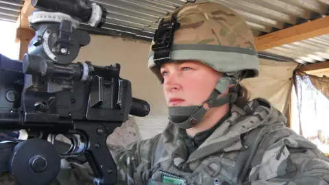 Nadine Askew wearing a camouflage military uniform and rounded helmet with a chin strap. She is sitting underneath a corrugated metal shelter, looking towards the left as she aims a machine gun.
