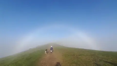 BBC Weather Watchers/Stormy D A person's shadow shows them taking a photo of another person walking a dog on the hills with blue sky around them.