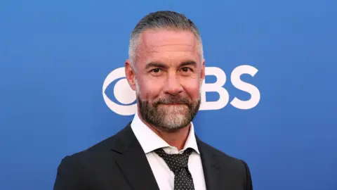 Actor Jay Harrington wearing a black suit with a white shirt and dark patterned tie. He is standing in front of a poster board which is blue and has the CBS logo on it.