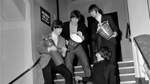 Getty Images Paul McCartney, John Lennon and Ringo Starr stand on the steps leading to the seats with George Harrison sitting down on them. They're sharing a joke and posing while holding a fire entinguisher and buckets.