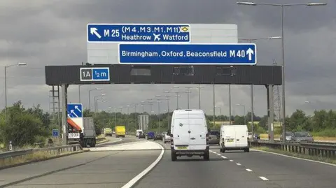 Vehicles in two lanes on the M40 travel under a large blue sign that directs them either to Heathrow and Watford or Birmingham, Oxford and Beaconsfield. The picture has been taken on a stormy day.