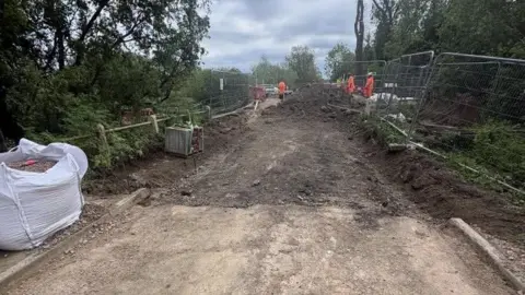North Northamptonshire Council Workers can be seen in the distance wearing orange clothing. In the foreground is a concrete road and piles of earth.
