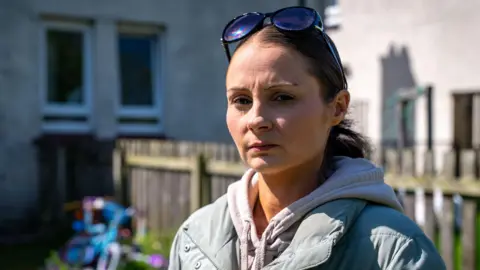 BBC Laura, in a grey puffer jacket and grey hoodie, is standing in a garden. A child's bike can be seen behind her.