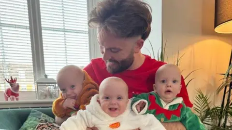 handout Jamie McEvoy grins as he looks at the baby triplets, who he holds together in his arms. They are on a sofa with a window behind.