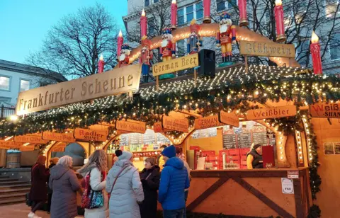 A stall selling alcohol and hot drinks at a Christmas market. The wooden stall is adorned with fake Christmas tree sprigs, golden fairylights, signs saying "Frankfurter Scheune", "Weissbier", "Radler Shandy". A group of people in warm winter coats and woolly hats stand in front of the stall