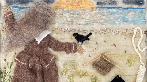 We Are Witch/Val Thomas Quilted panel made from fabric depicts a woman with grey hair in a long grey dress or gown, with a crow-like bird on her right hand. She appears to be looking out to sea where the sun is going down. Knitting needles and knitting can be seen, along with the top part of a hangman's rope.