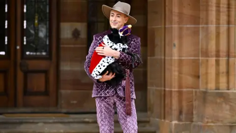 Getty Images Alan Cumming, a man, in a purple diamond quilted suit, a grey suede cowboy hot, stood central to the photo infront of a building and he is smiling looking to the left side of the photo. He is cradling a black dog in his arms, the dog is wearing a crown and has a royal cape on. 