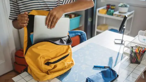 Child's arm putting grey laptop in yellow school bag on a desk, which has a blue pencil case on it.