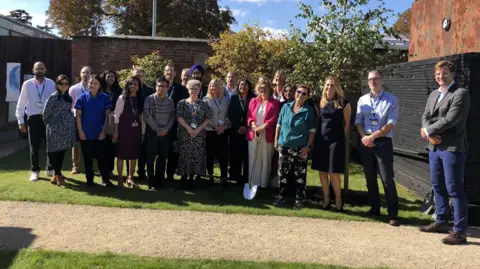 BBC A group of healthcare professionals and other representatives gathered around the new tree at Glenfield Hospital