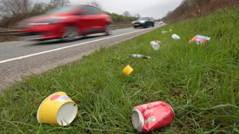 Close-up of roadside verge showing litter, including a paper cup and a Coke can, while traffic (blurred) passes on the road nearby