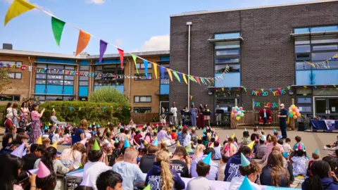 Wood Farm Primary School A crowd of school children, all wearing colourful party hats, are sat below bunting and in front of a large school building.