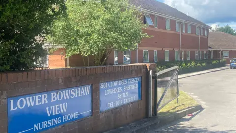 Lower Bowshaw View from its front gate. The main building is made from red brick, and the name of the home is printed on a blue sign next to the gate.