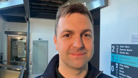 A close-up image of Conchúr Ó Muadaigh. He has short brown hair and is smiling with his mouth closed at the camera. He is stood in front of white walls with a floor directory on his right hand side.