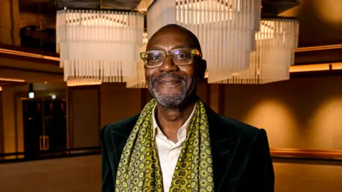 A man with short dark hair, a white shirt and dark jacket with a patterned yellow and black scarf around his neck and brown walls behind him