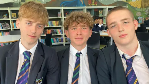Three people seated close together in the foreground, facing directly toward the camera. All three individuals are wearing matching dark blazers, white shirts, and striped ties with diagonal bands in multiple colors, suggesting a school uniform. The person on the left has light-colored, slightly tousled hair, while the person in the middle has short, curly hair, and the person on the right has very short hair.
They are sitting in what appears to be a library or study area. Behind them, there are shelves filled with a variety of books arranged upright and in rows. The shelves are white and evenly spaced, creating a structured and organized background.