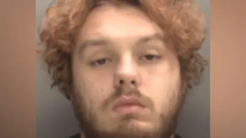 West Midlands Police Gabis pictured in his police mugshot. He has a black expression on his face. He has light brown curly hair and brown facial hair.