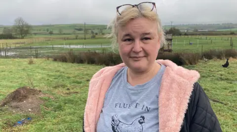 A woman with blonde hair tied back and glasses resting on her head is looking into the camera with a serious expression. She has a nose ring and a few other piercings on her face and is wearing a grey top with a chicken on it and a black coat with a pink fleece lining. She is stood in a field where two livestock animals can be seen in the background.