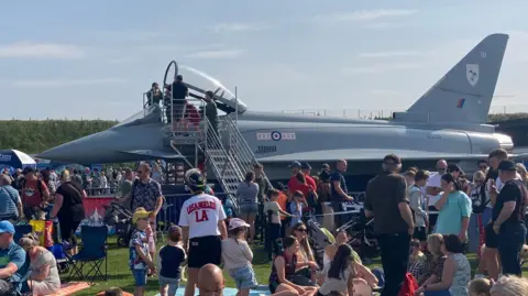 Some people stand around the seating area of a grey jet. There are a lot of people standing on grass around the jet.