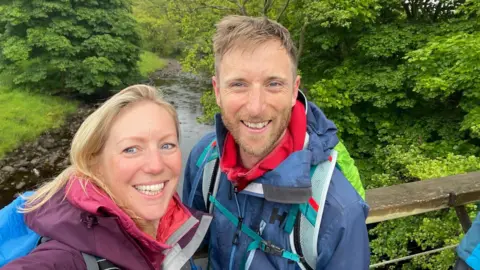 MY NAME’5 DODDIE FOUNDATION Luke Hames-Brown is on the right, wearing a waterproof coat and carrying a rucksack; Kate, his wife, is wearing a purple coat and is also carrying a rucksack