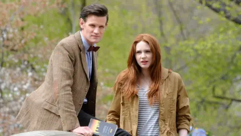 Getty Images Matt Smith and Karen Gillan filming on location for "Doctor Who" in Central Park on April 11, 2012 in New York City. Matt wears a blue shirt with a dark red bow tie and a checked brown jacket. Karen wears a blue and white striped top and a brown anorak. They are outside in what appears to be a park.