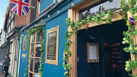 The outside of the pub which is painted blue and has a floral garland around the door. There are signs on the outside wall and inside the entrance. through the door, rows of drinks can be seen. The building has a bay window, which is also decorated, and there is a flag above the tap room. Further along the high street, a woman is standing underneath a black and white building.