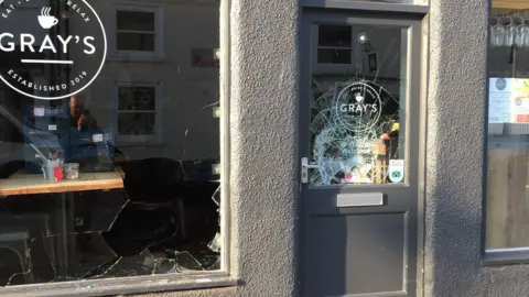 Gray's of Rothwell A smashed window and a broken grey door with a circular Gray's logo.