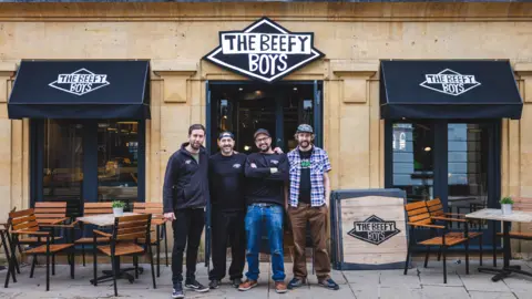 Four men stood outside a shop called "Beefy Boys" in black and white text. They are wearing dark tops and jeans. There are two tables surrounded by wooden chairs either side of them.