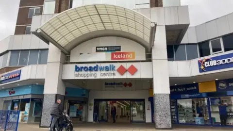 Broadwalk Shopping Centre in Knowle, Bristol. There are signs for businesses inside and beside the main entrance of the shopping centre. There is a pedestrian stood outside the entrance.