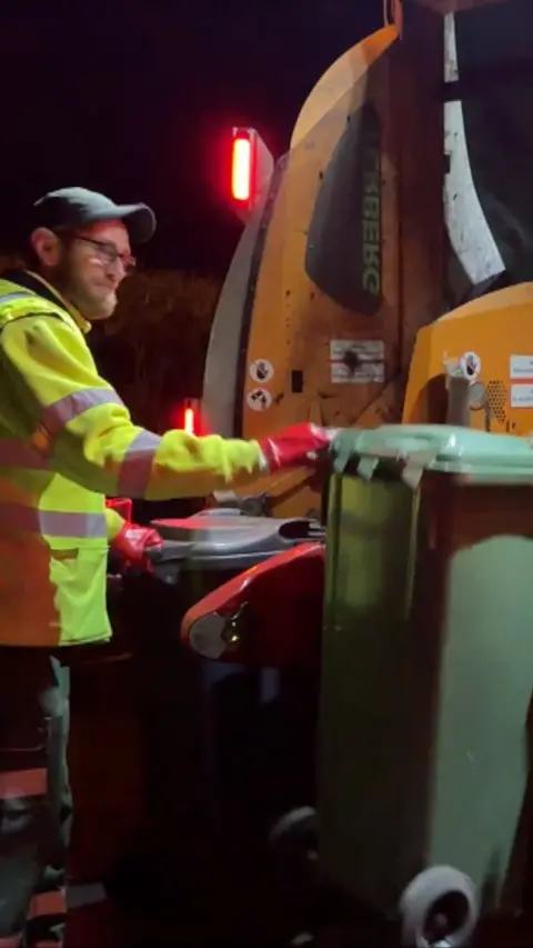 A man in hi vis jacket and weraing a cap emptying a large plastic bin into a bin truck 