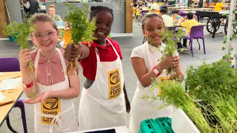 Three schoolgirls, each holding a bunch of small carrots, stand in front of a table on which an open box of carrots sits. 