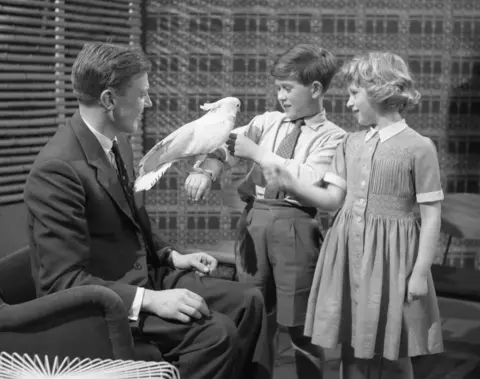 A black and white image of two children and man in a suit. The boy holds a white cockatoo on his arm whilst all watch on. 